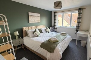 A bedroom with a bed, a bedside table, a lamp, a window with curtains, and a view of buildings outside.