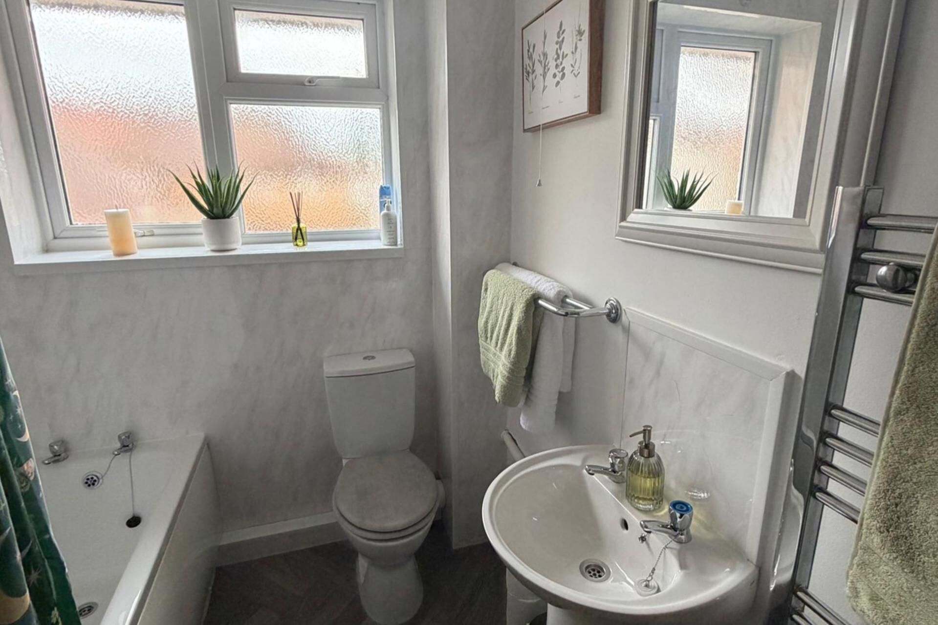 Une salle de bain avec des toilettes, un lavabo et une baignoire. Il y a deux fenêtres, et des plantes sont posées sur le rebord de chaque fenêtre.