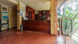 Uma área de recepção com balcão de madeira, piso de azulejos e vista para palmeiras através de uma porta de vidro.