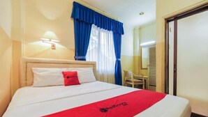 Quarto de hotel com uma cama, uma mesa, uma cadeira, uma janela com cortinas e uma porta.