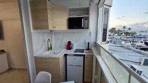 A compact kitchen with a microwave, sink, and small refrigerator. A dining table with placemats and a cup is visible. The room has a balcony with a view of a marina and boats. - Charming studio overlooking Saint-François marina, golf course, lagoon & beach on foot. (Saint-François)