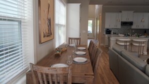 Un comedor con una mesa de madera dispuesta para seis personas, rodeada de sillas de madera. Se ve una cocina al fondo con armarios blancos y una isla integrada con taburetes de bar.