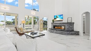 A spacious living room with a fireplace, a television, and a large window overlooking a scenic view. - Seashell Paradise: 5BR w/ Pool, Spa on Sanibel! (Sanibel)