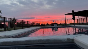 Une piscine entourée d’une clôture en métal, un espace abrité et un coucher de soleil en arrière-plan.