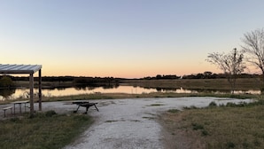 Un sentier de gravier mène à un espace de pique-nique équipé d’un abri et de bancs, situé au bord d’un lac paisible, au moment du coucher du soleil.