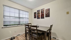 A dining area with a table and chairs, a window with blinds, and three wall-mounted artworks.