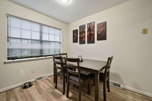 Ein Essbereich mit Tisch und Stühlen, einem Fenster mit Jalousien sowie drei an der Wand befestigten Kunstwerken.