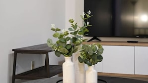 A living room with a round table, three white vases with plants, a remote control, and a television on a stand.