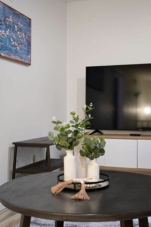 Ein Wohnzimmer mit einem runden Tisch, drei weißen Vasen mit Pflanzen, einer Fernbedienung und einem Fernseher auf einem Standfuß.