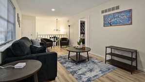 A living room with a sofa, a coffee table, and a TV stand. There is a painting on the wall and a window with blinds.