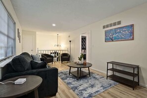 Ein Wohnzimmer mit einer Couch, einem Couchtisch und einer TV-Konsole. An der Wand hängt ein Gemälde und ein Fenster mit Jalousien.
