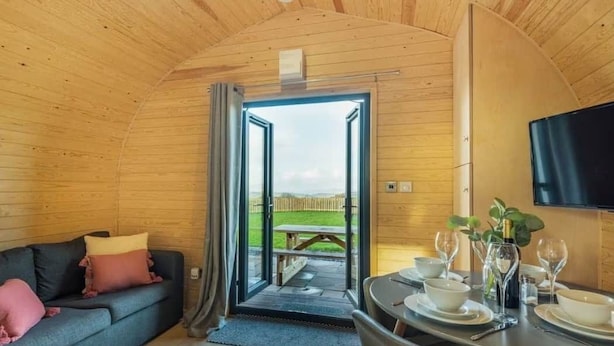 Ein gemütlicher Raum mit Holzbalkendecke, einer Couch mit Kissen, einem Esstisch mit Tellern, Gläsern und einer Flasche sowie einem an der Wand befestigten Fernseher.