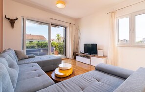 Ein modernes Wohnzimmer mit einer Sofagarnitur, einem Fernseher und einem Balkon mit Blick auf Häuser und Grünflächen.