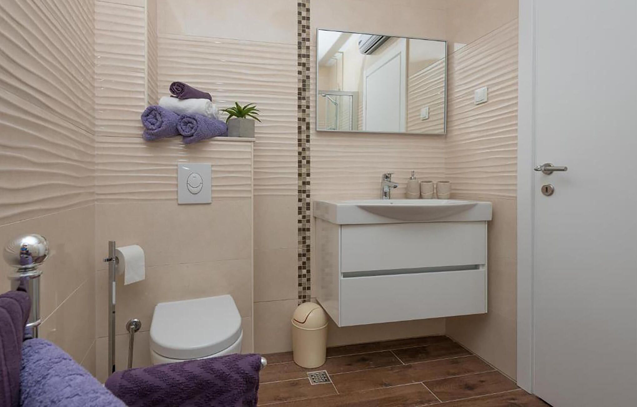 A bathroom with a toilet, sink, and mirror. There are towels and a trash can.