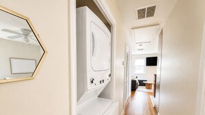 Un lavadero con lavadora y secadora, un ventilador de techo, y un pasillo que conduce a una sala de estar con sofá y televisor.