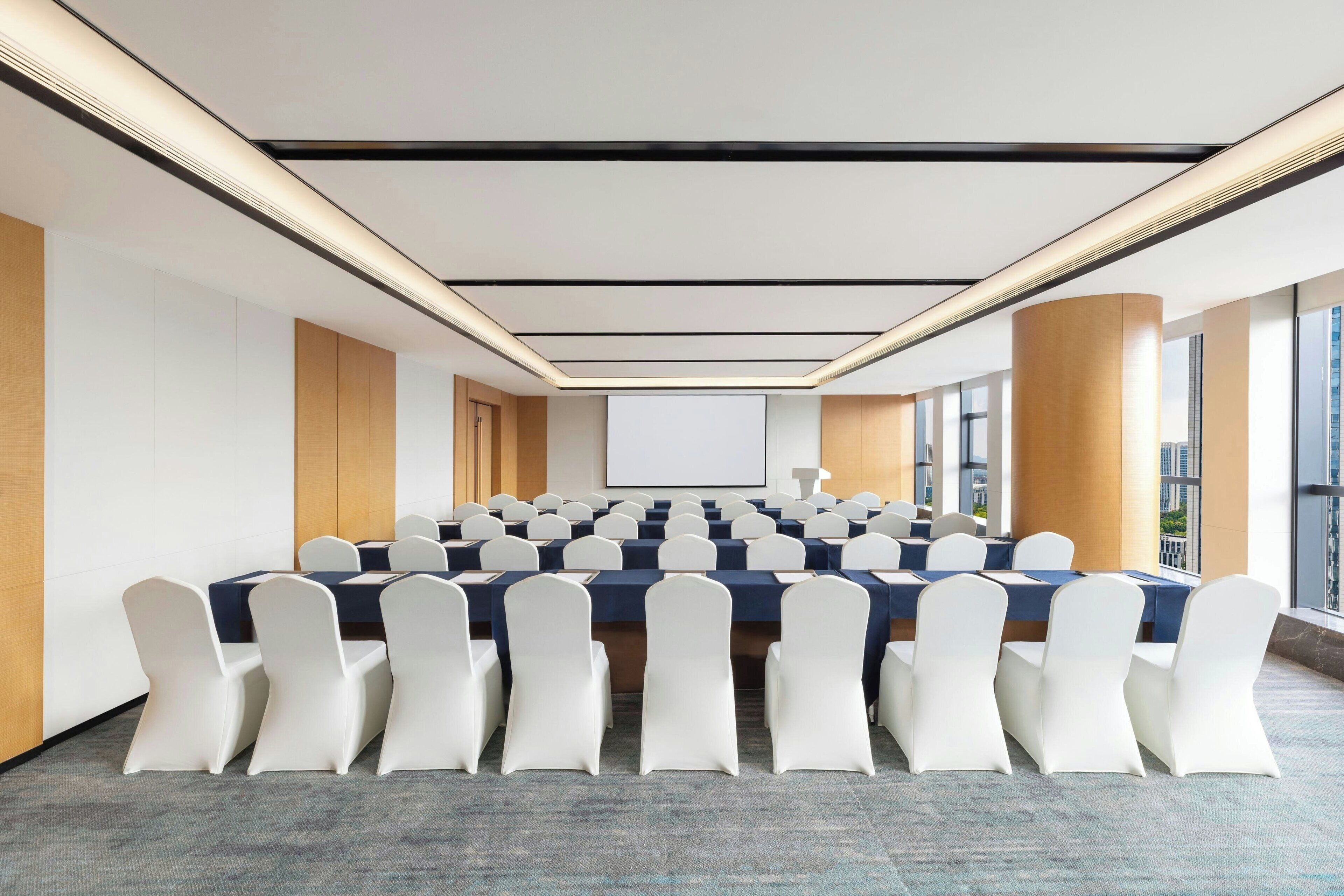 A conference room with a long table, white chairs, and a projector screen.