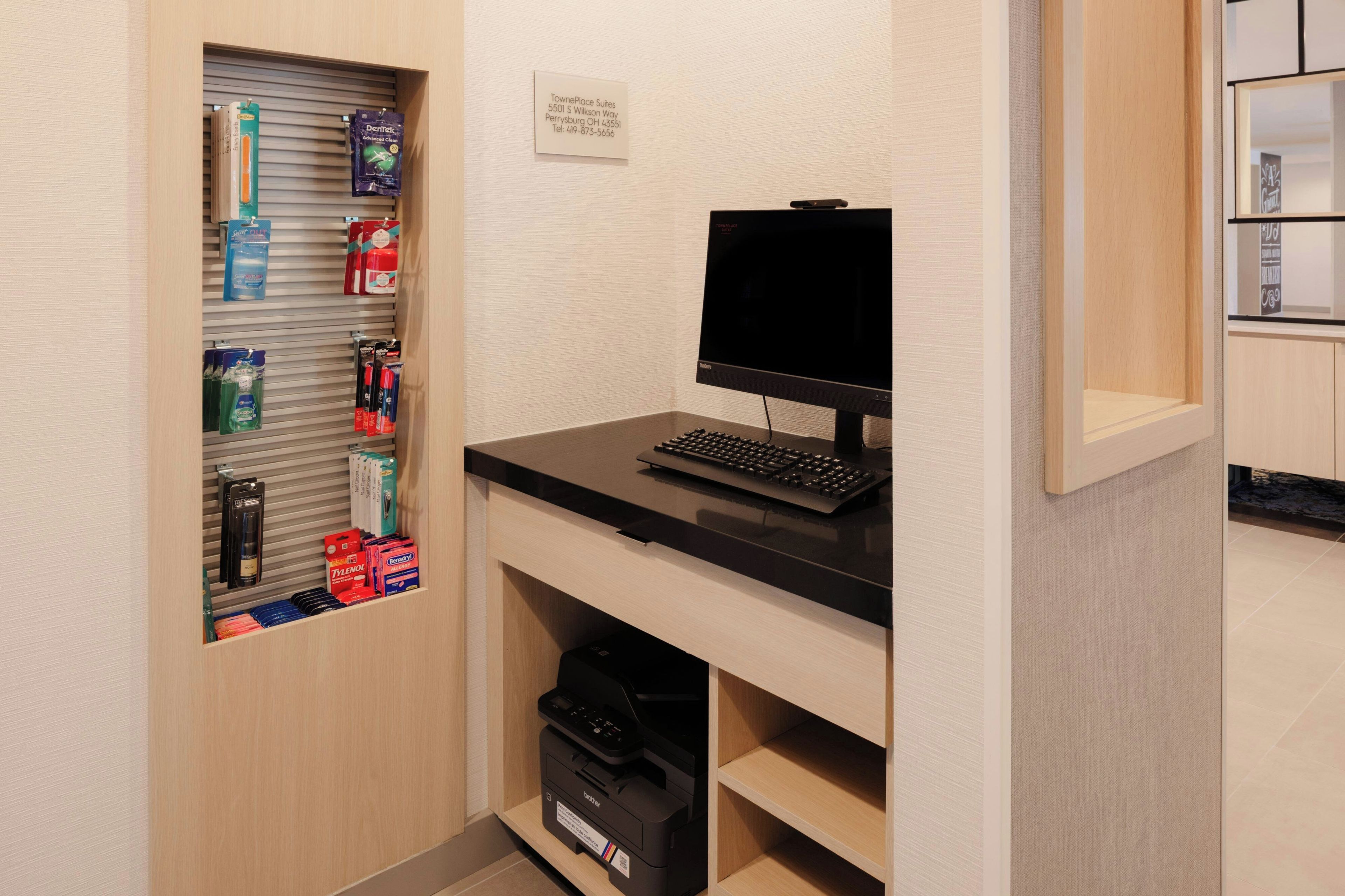 A hotel room with a desk, computer, printer, and a shelf with toiletries.