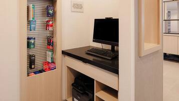 A hotel room with a desk, computer, printer, and a shelf with toiletries.