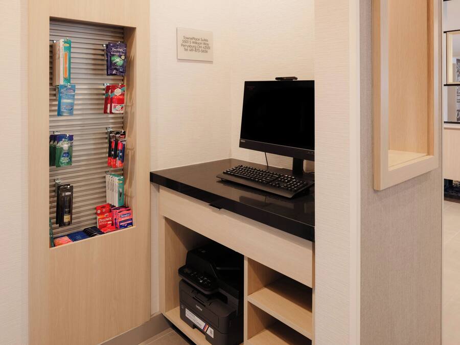 A hotel room with a desk, computer, printer, and a shelf with toiletries.