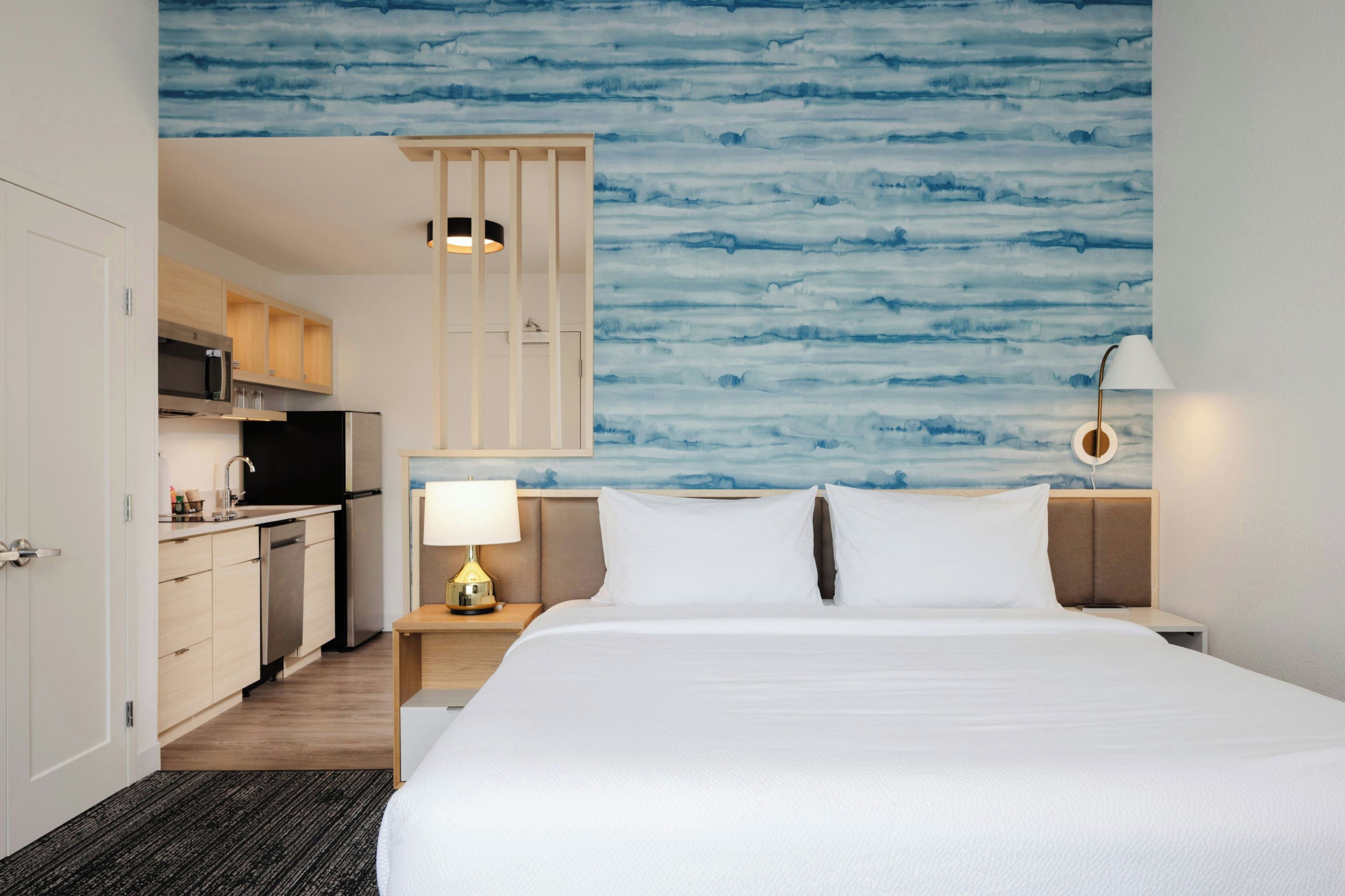 A hotel room with a bed, a nightstand, a lamp, a desk with a chair, a kitchenette, and a bathroom.