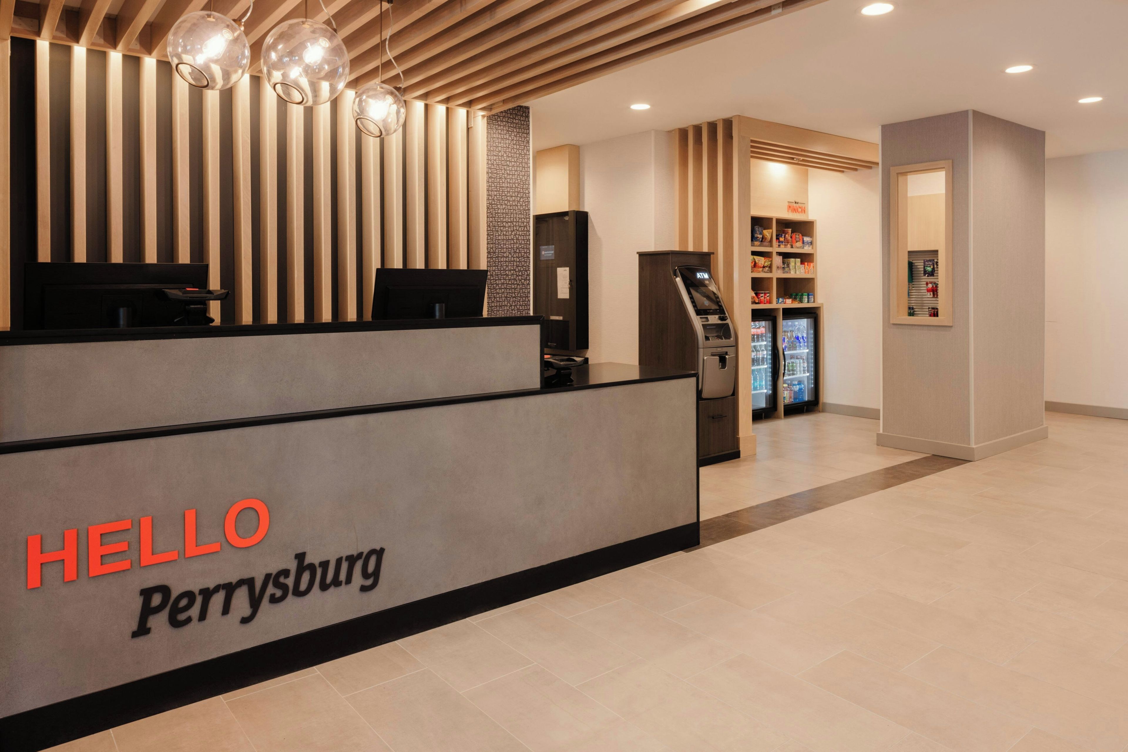 A reception desk with a 'HELLO PERRYSBURG' sign, a cash register, a refrigerator stocked with beverages, and a snack shelf.