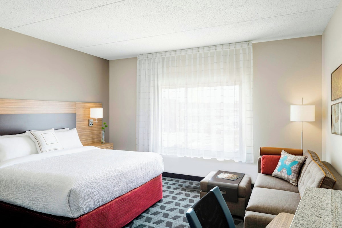 Una habitación de hotel con una cama grande, un sofá, una mesita y una ventana con cortinas traslúcidas.