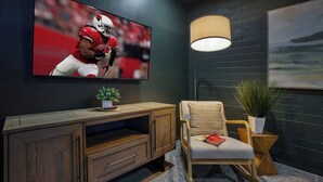 A living room with a television displaying a sports game, a wooden entertainment unit, a comfortable chair, a floor lamp, and a painting on the wall. - Downtown Gilbert • Hot Tub • Glow Golf • Sleeps 12 (Gilbert)