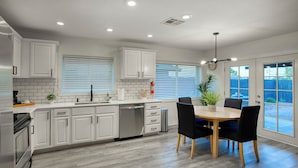 A modern kitchen with white cabinetry, a built-in dishwasher, and a dining area with a wooden table and black chairs. - Downtown Gilbert • Hot Tub • Glow Golf • Sleeps 12 (Gilbert)