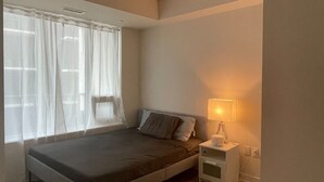 A bedroom with a bed, a nightstand with a lamp, and a window with sheer curtains.