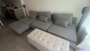 A modern living room with a grey sectional sofa, a white ottoman, and a dining area with a small table and chairs.