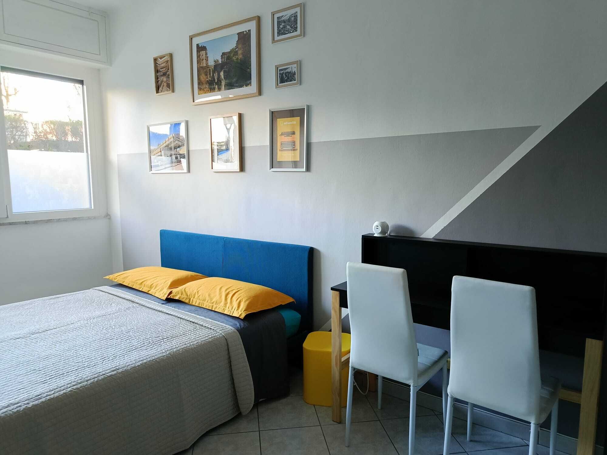 A bedroom with a bed, a desk, two chairs, a window, and several framed pictures on the wall.