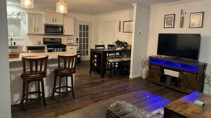 Una cocina moderna con armarios blancos, un comedor con mesa y sillas de madera, y una sala de estar con sofá de cuero y televisor.