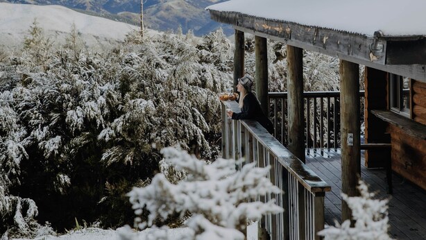Un bâtiment en bois, avec un toit enneigé et un balcon donnant sur un paysage hivernal, avec des montagnes à l’horizon.