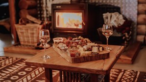 Une table en bois, sur laquelle sont disposés une planche de charcuterie et deux verres de vin, dans une pièce chaleureuse avec une cheminée et un téléviseur en arrière-plan.