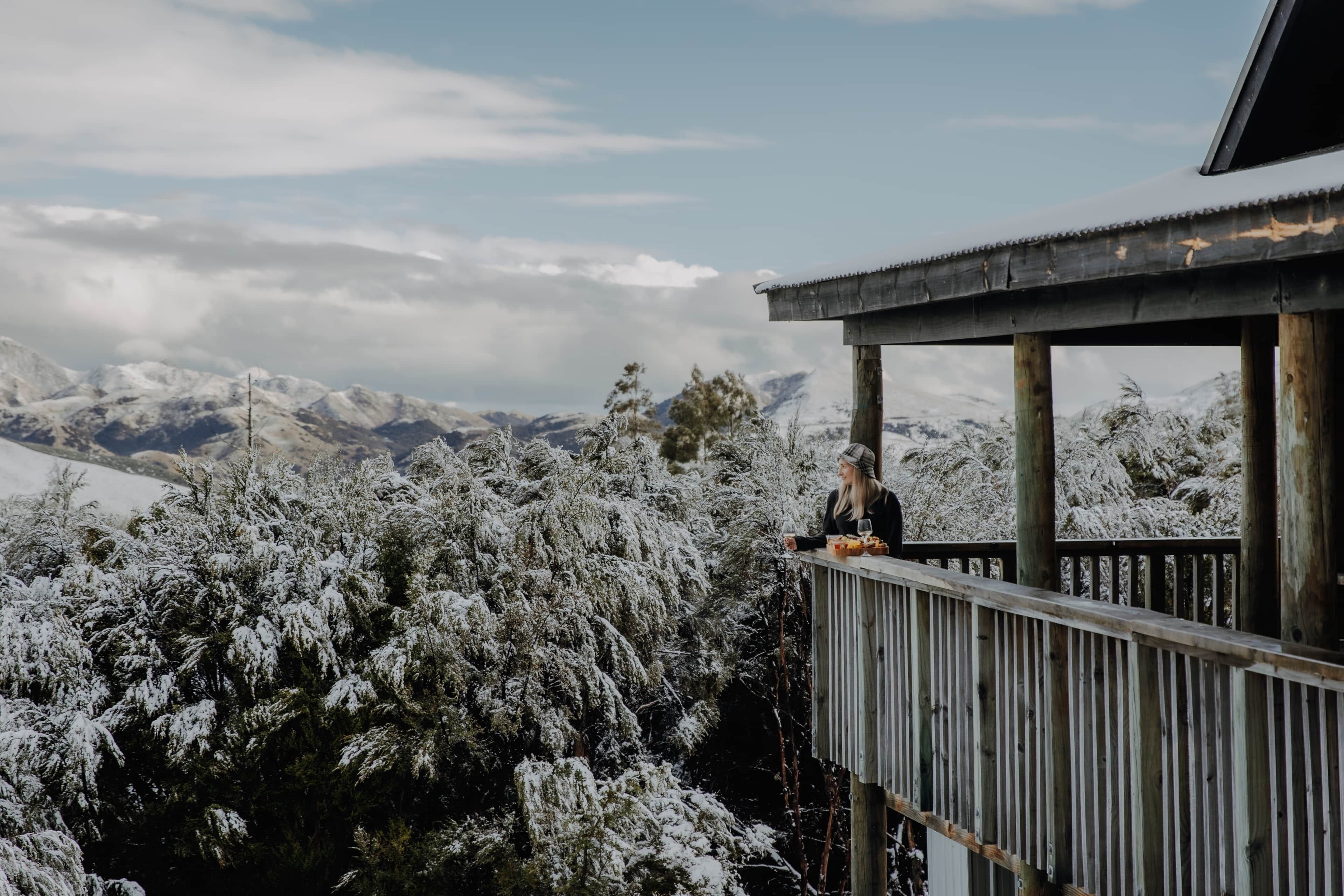 Une personne assise sur un balcon, avec vue sur des arbres et des montagnes couverts de neige.