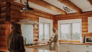 L’intérieur d’un chalet rustique, avec un plafond en bois, un coin cuisine équipé d’un poêle et une fenêtre donnant sur une forêt.
