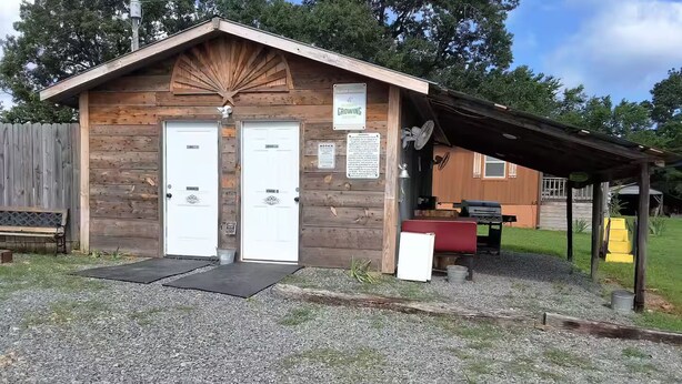 A wooden cabin with two white doors, a covered outdoor area, and a small grill. - Arabian Cabin (Heavener)