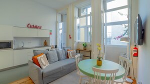 A compact living space with a kitchenette, a grey sofa with orange cushions, a round green table with white chairs, and a large window providing natural light. - VIVA República Serviced Apartment 0T (Porto)