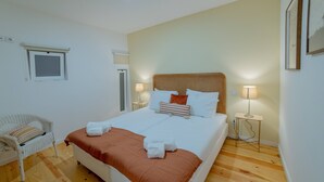 A neatly arranged bedroom with a large bed, two bedside tables with lamps, a wicker chair, and a window with blinds. - VIVA República Serviced Apartment 0T (Porto)