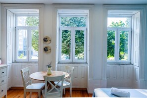 A room with a wooden floor, white walls, and a window with a view of greenery. There is a bed with a white blanket and a small table with a vase. - VIVA República Serviced Apartment 1F (Porto)