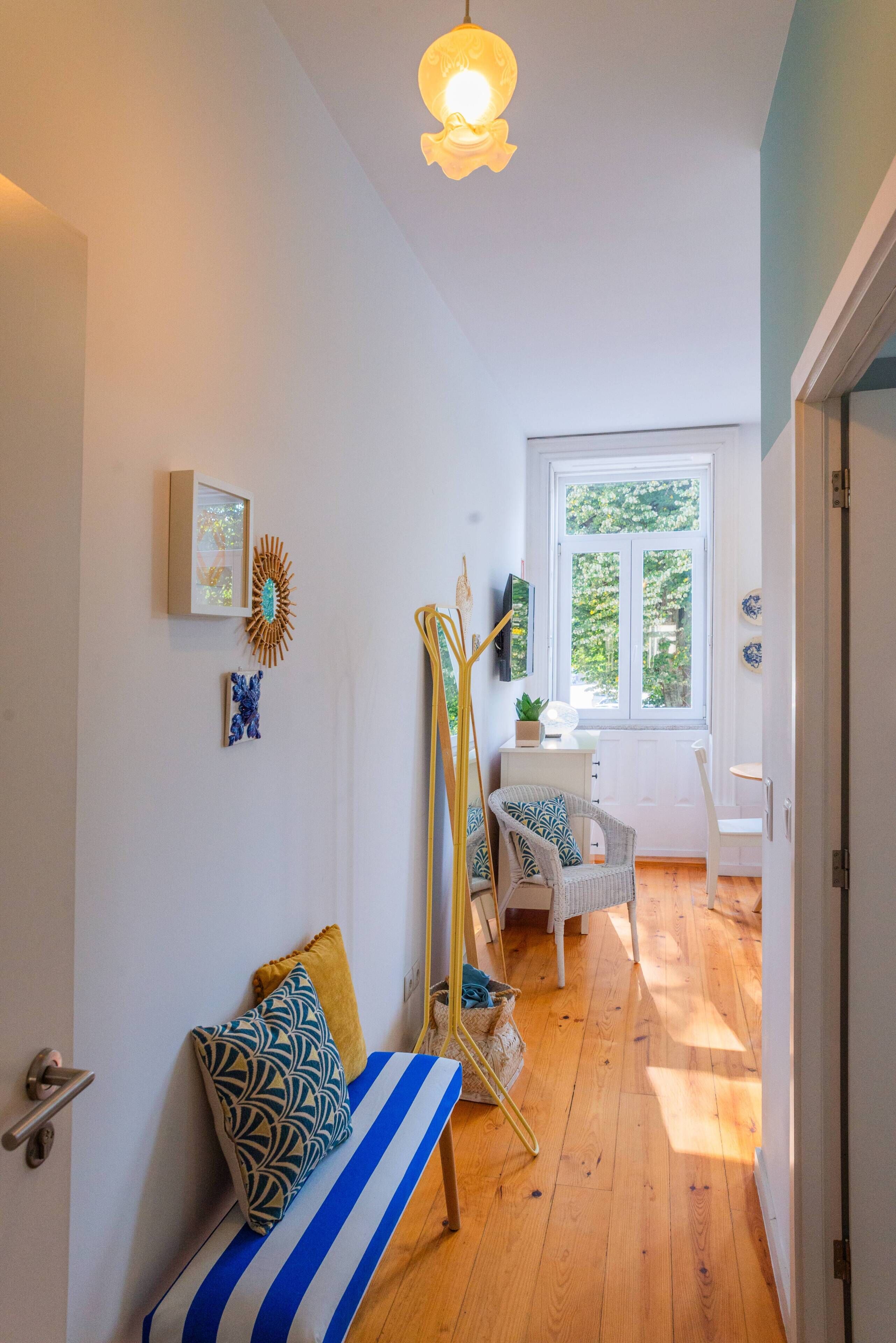 A bright hallway with a wooden floor, a bench with patterned cushions, a standing lamp, and a window with a view of greenery.
