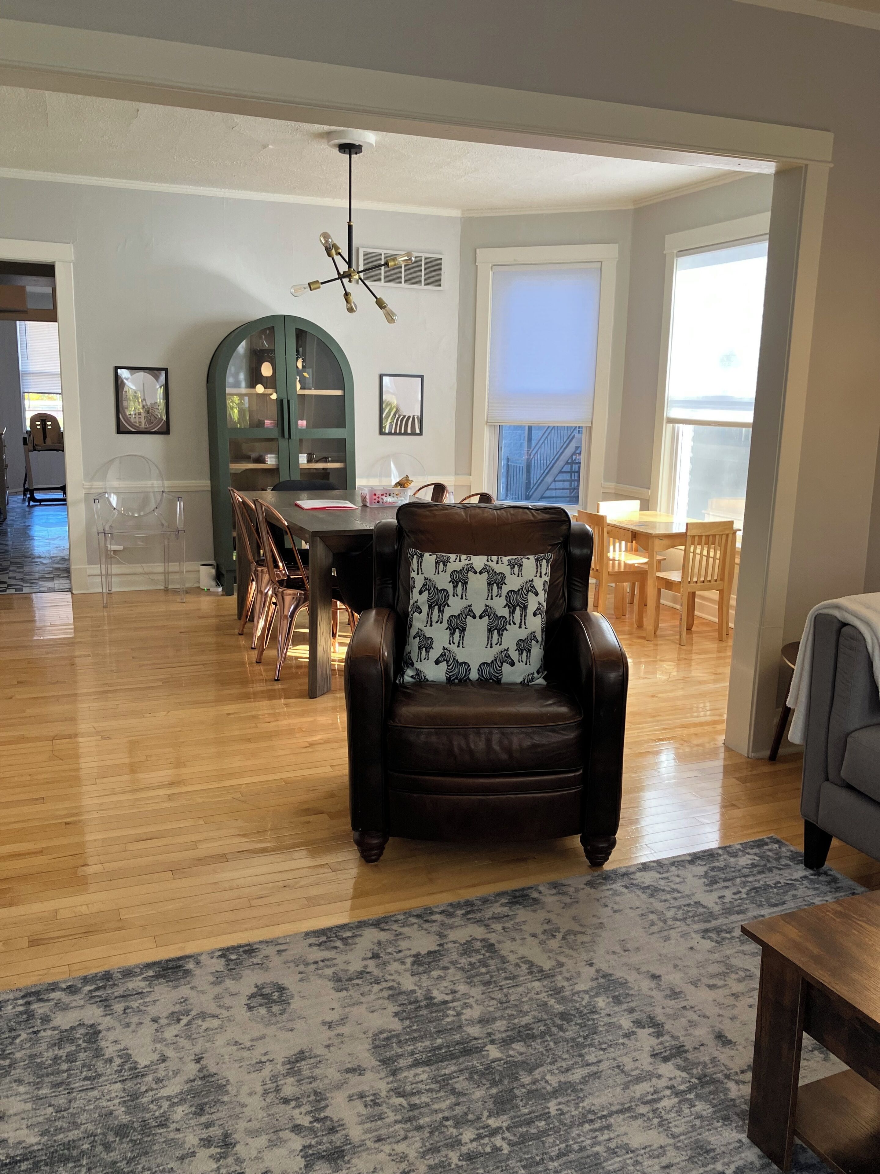 A spacious living room with a wooden floor, a leather armchair with a zebra print cushion, a dining area with wooden chairs and a green cabinet, and a large window providing natural light.