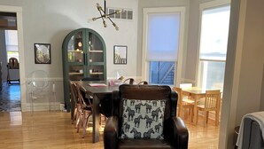 Un vaste salon doté d’un plancher en bois, d’un fauteuil en cuir agrémenté d’un coussin à motifs zébrés, d’un coin repas avec des chaises en bois et un buffet vert, ainsi qu’une grande fenêtre laissant entrer la lumière naturelle.