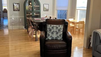 A spacious living room with a wooden floor, a leather armchair with a zebra print cushion, a dining area with wooden chairs and a green cabinet, and a large window providing natural light.