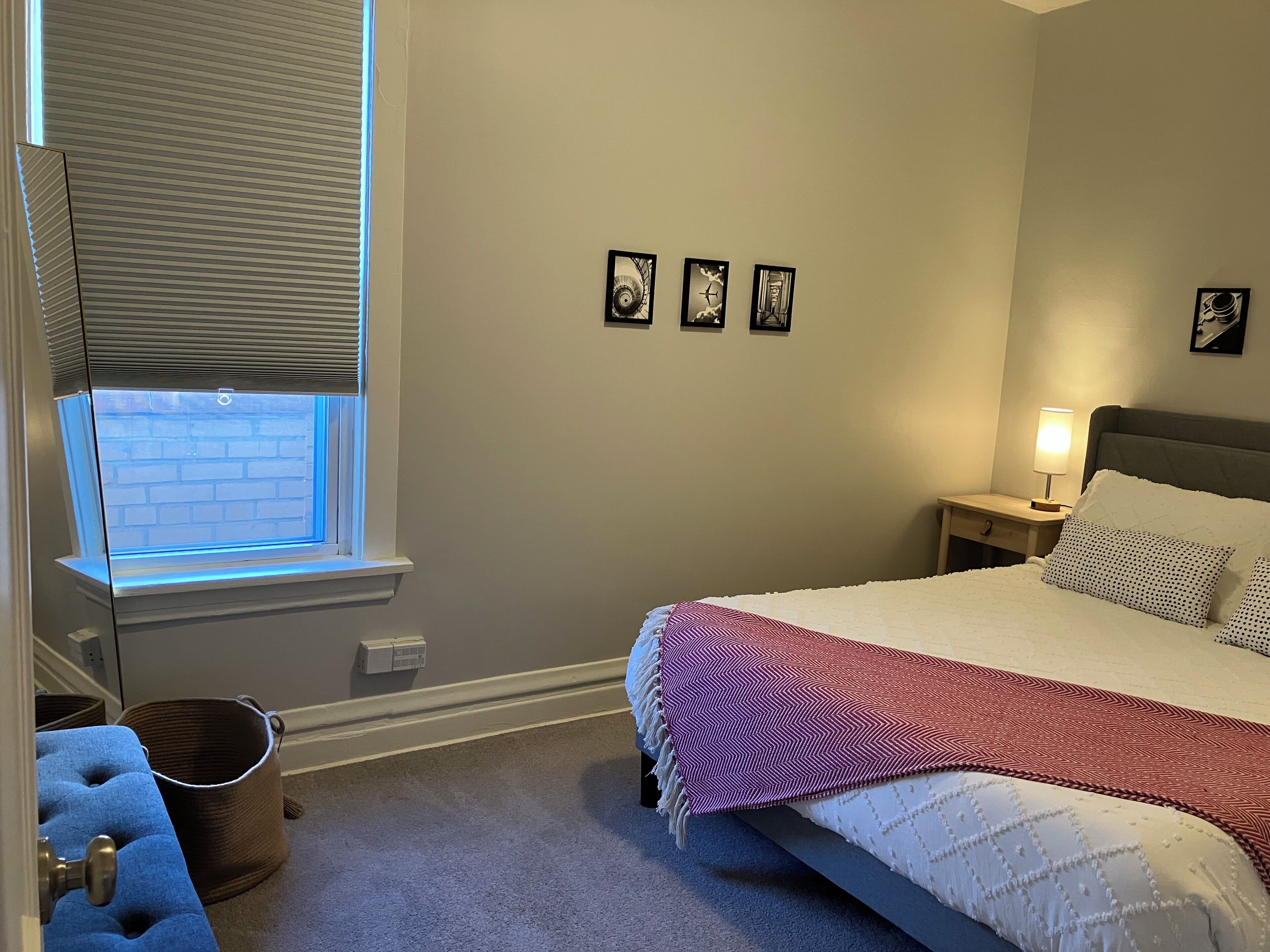 Une chambre à coucher comprenant un lit, une table de chevet, une lampe, une fenêtre avec des stores, une étagère fixée au mur et un petit panier.