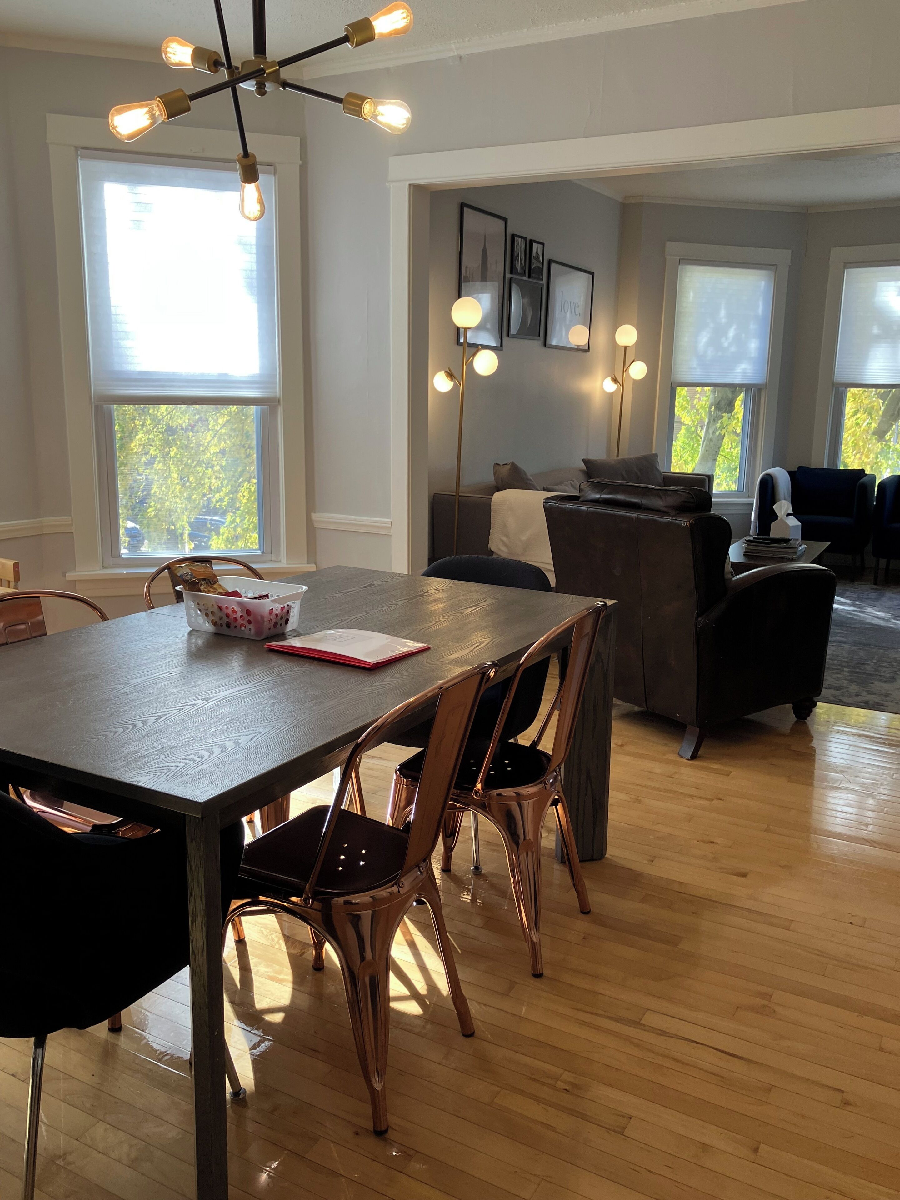 A dining area with a wooden table, four chairs, and a chandelier. There are large windows with blinds, a sofa, and a coffee table in the background.
