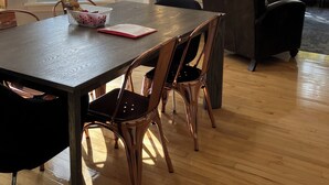 Un coin repas avec une table en bois, quatre chaises et un lustre. De grandes fenêtres équipées de stores, un canapé et une table basse se trouvent à l’arrière-plan.