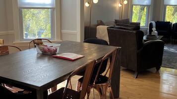 A dining area with a wooden table, four chairs, and a chandelier. There are large windows with blinds, a sofa, and a coffee table in the background.