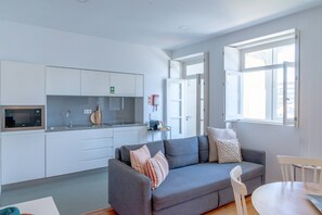 A modern living room with a grey sofa, white chairs, and a small table. A kitchen with white cabinets and a microwave is visible in the background. - VIVA República Serviced Apartment 2T (Porto)
