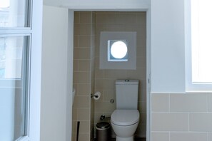 A small, narrow bathroom with a toilet, a trash bin, and a window with a circular skylight. - VIVA República Serviced Apartment 2T (Porto)